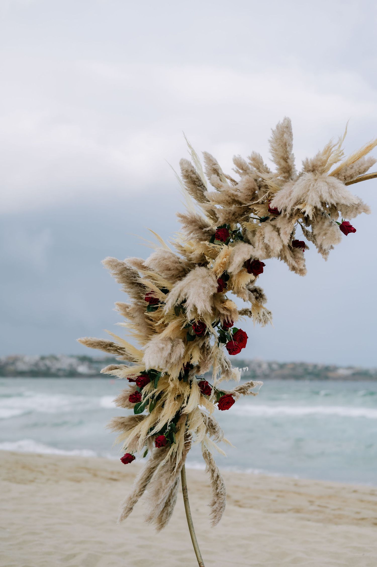 Dried pampas grass and red flower arrangement against ocean backdrop.