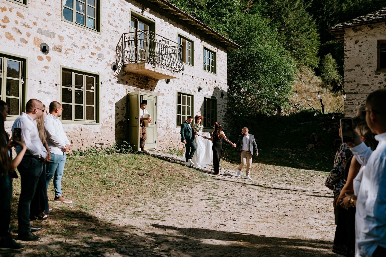Guests celebrate at an outdoor wedding reception near a traditional stone building.