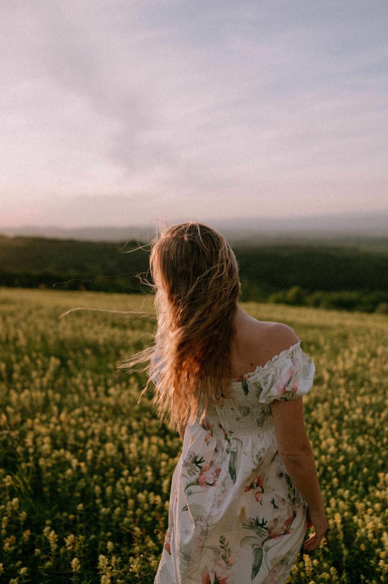 Person in floral dress with windswept hair in blooming yellow field.