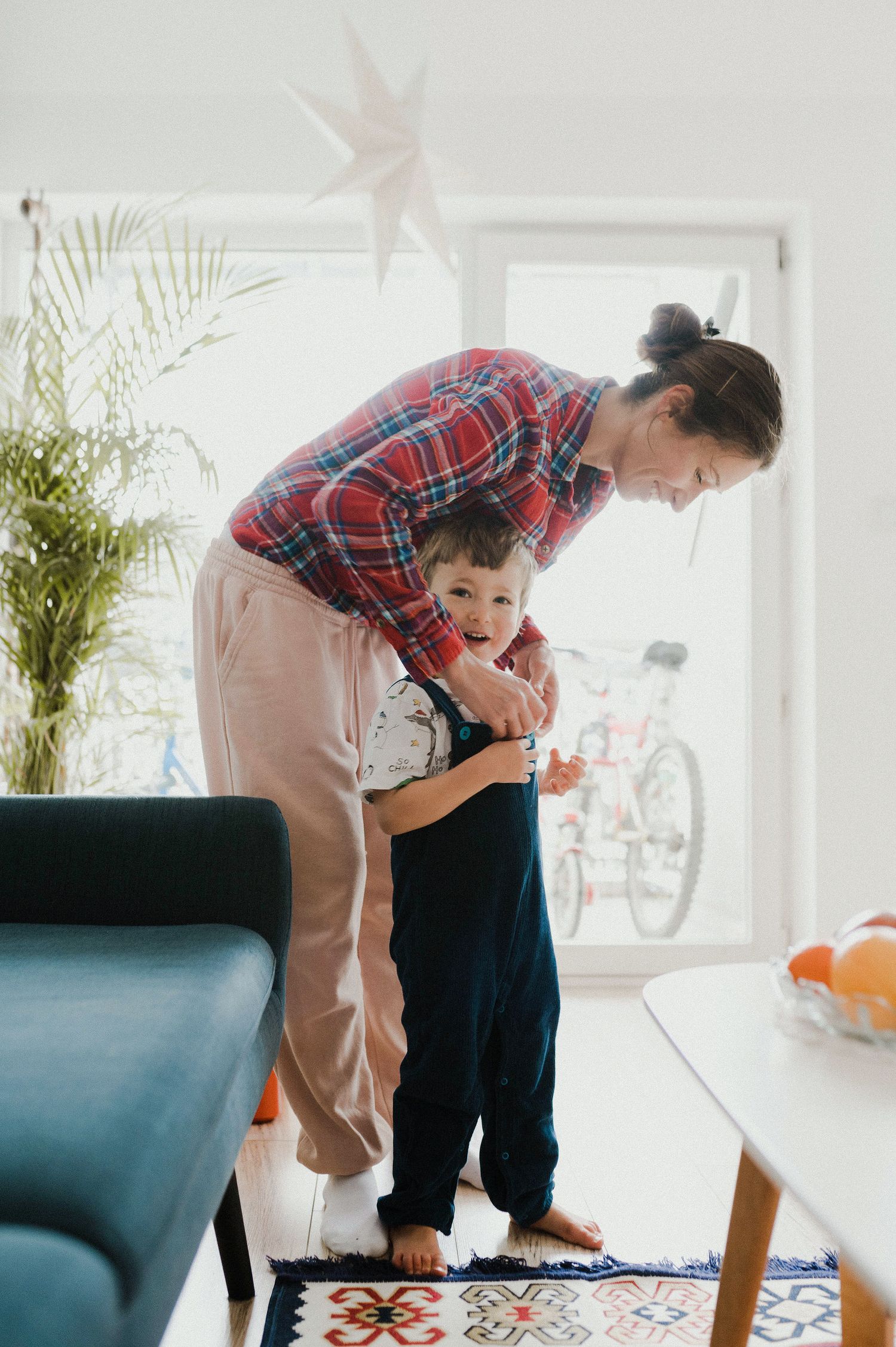 Parent in plaid shirt and khakis bends down to help young child in living room with large windows and plants.