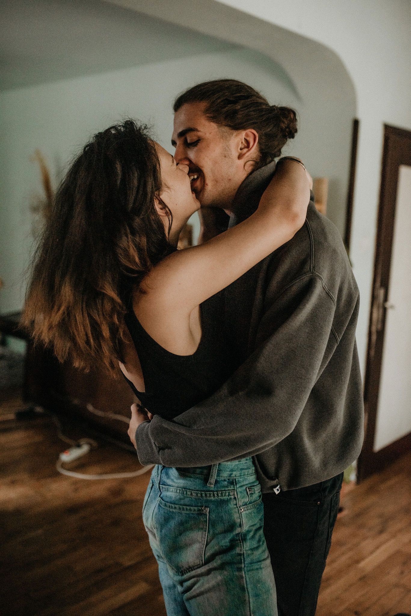 A romantic couple embraces and kisses in a dimly lit hallway with wooden floors and white walls.