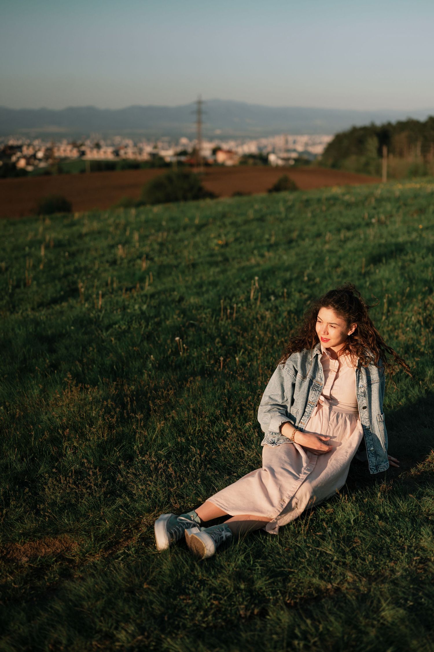 A person in light clothing relaxes on a grassy hillside overlooking distant mountains during golden hour.