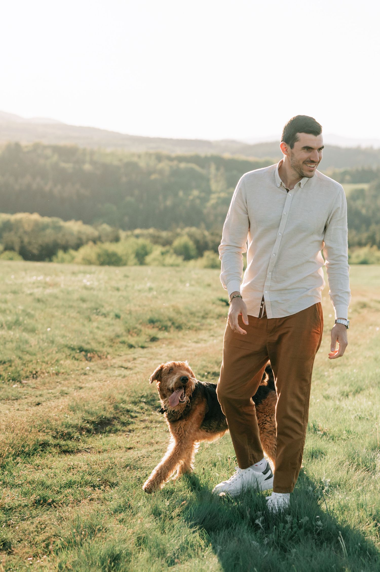 Person in white shirt and brown pants walking with an Airedale Terrier dog in a scenic mountain meadow at sunset.