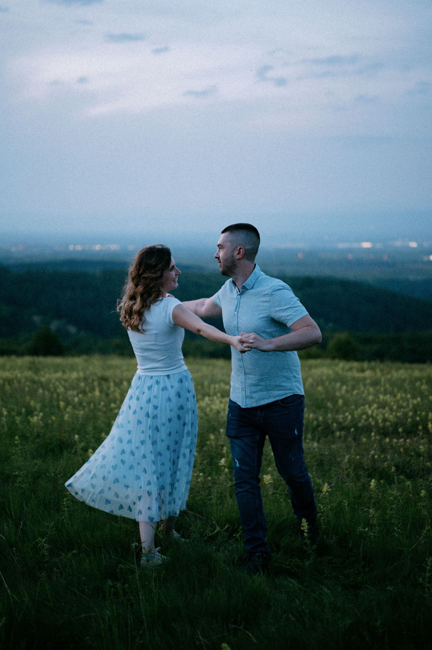A couple dances playfully in a grassy field with mountains in the background.