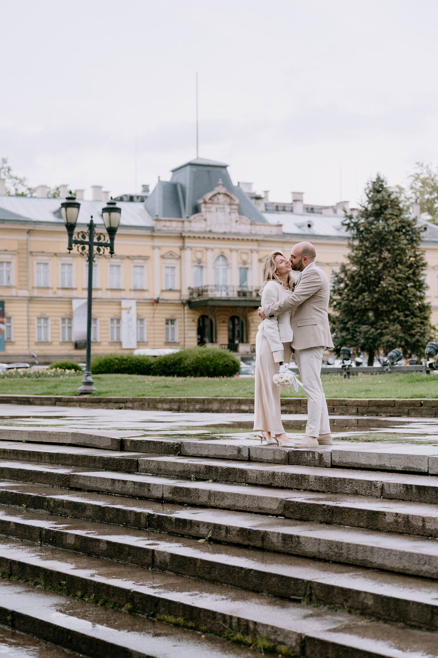Two people in light colored outfits share an intimate moment on stone steps near a historic building.