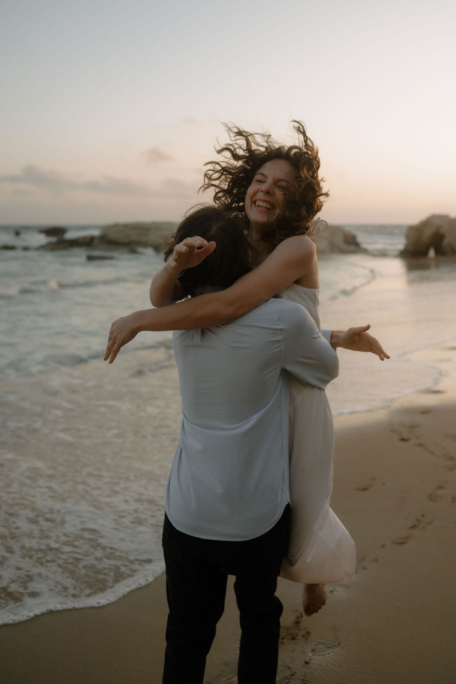 Couple embracing on a beach at sunset, woman laughing with hair blowing in the wind near ocean waves.