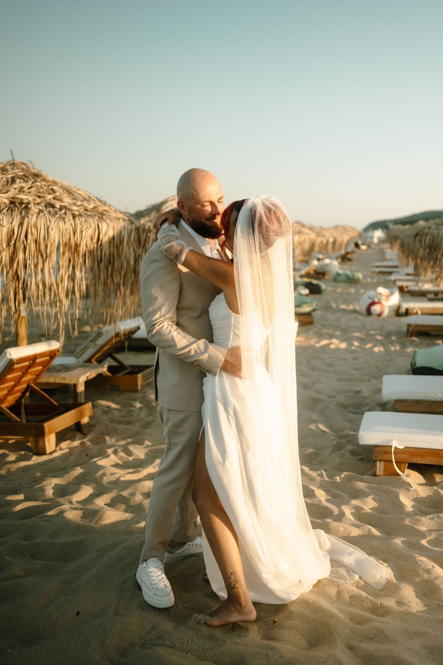 A tender moment between newlyweds on a beach with thatched umbrellas and lounge chairs in the background.