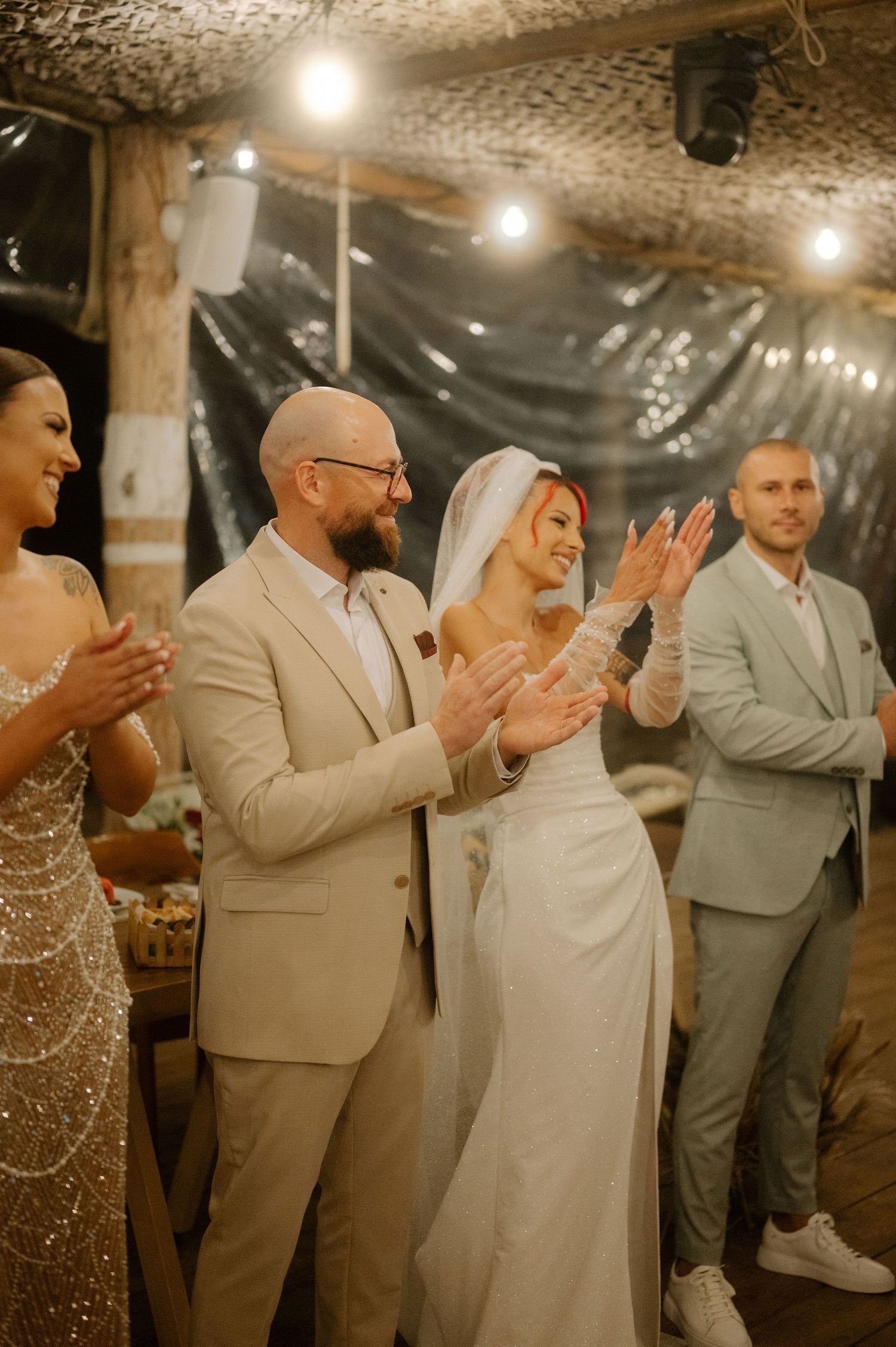 Wedding guests in formal attire clapping and celebrating during an evening reception under string lights.
