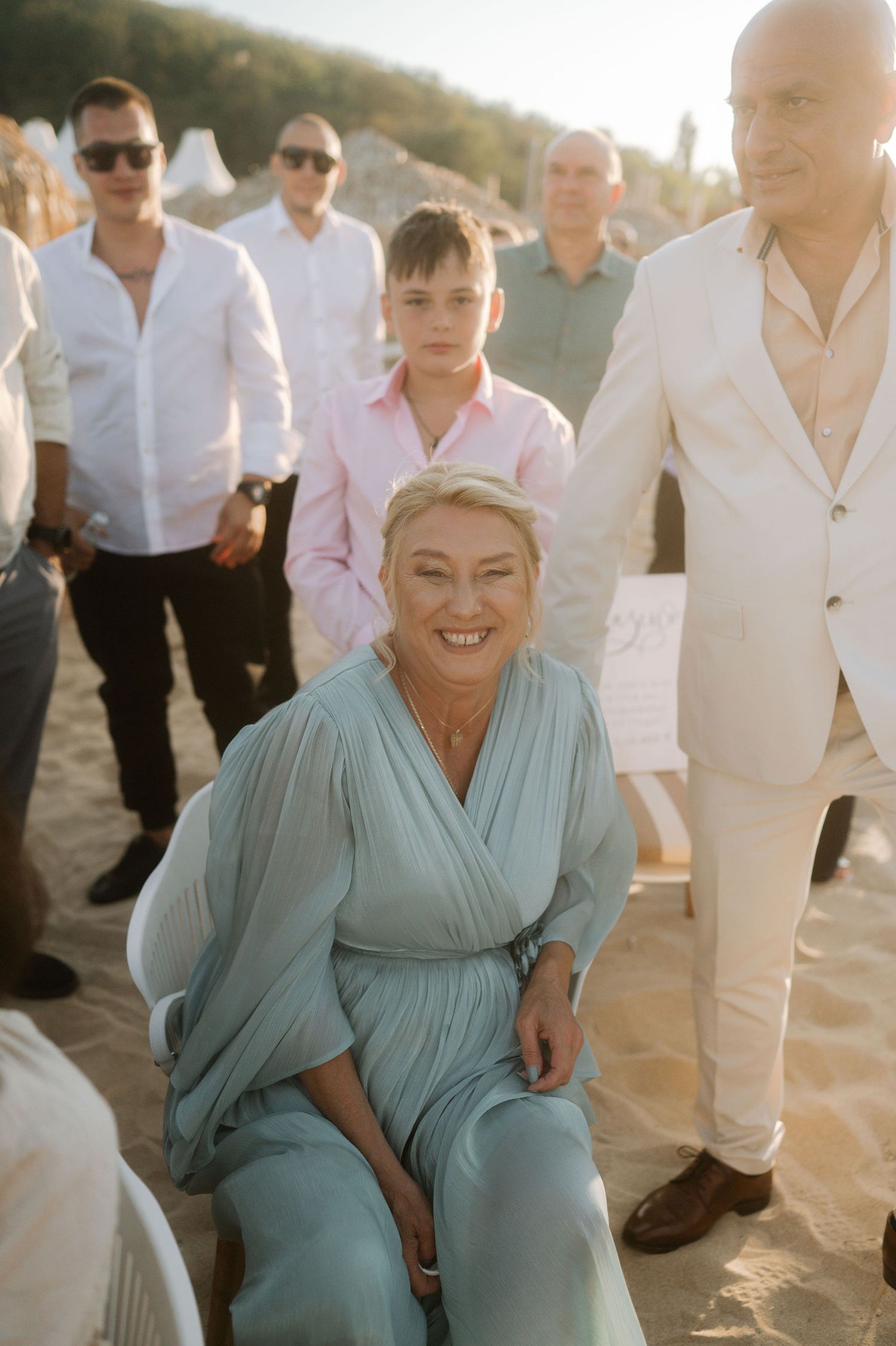 Wedding party members in white and blue attire laugh together on a sunny beach at sunset.