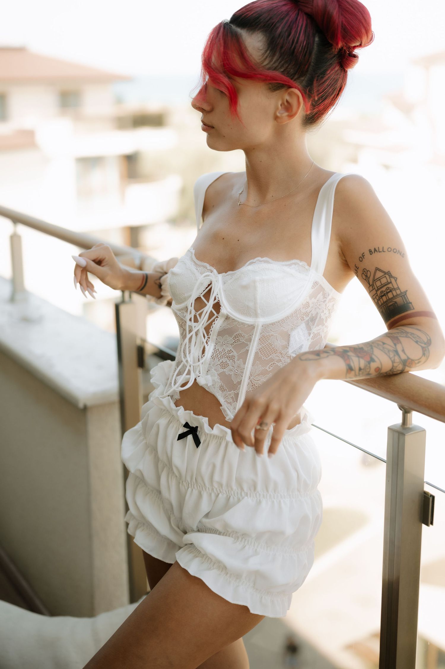 A person with red hair wearing a white corset top and shorts stands on a balcony with visible arm tattoos.