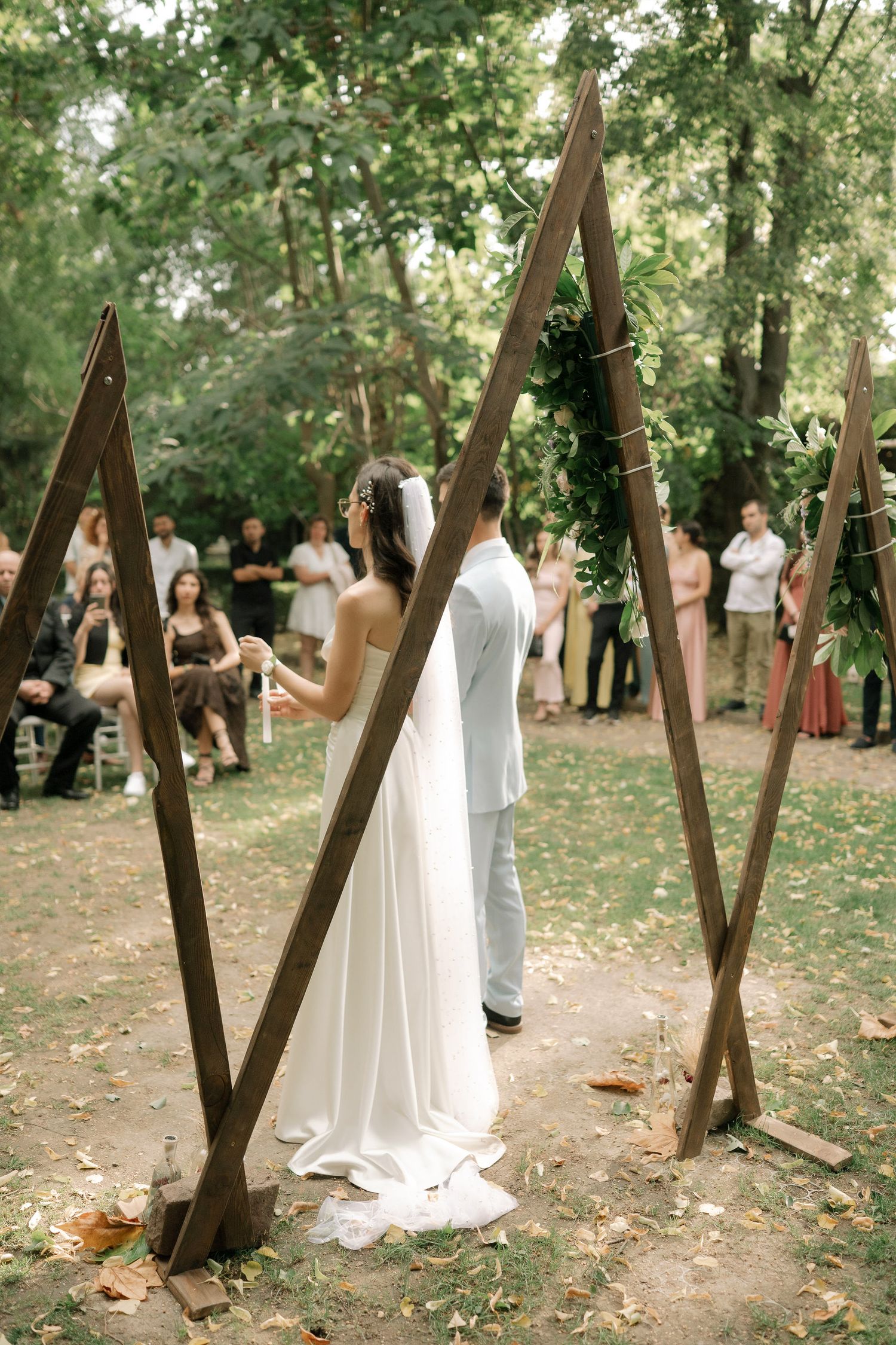 A couple in wedding attire stands among trees in a wooded outdoor setting during their ceremony.