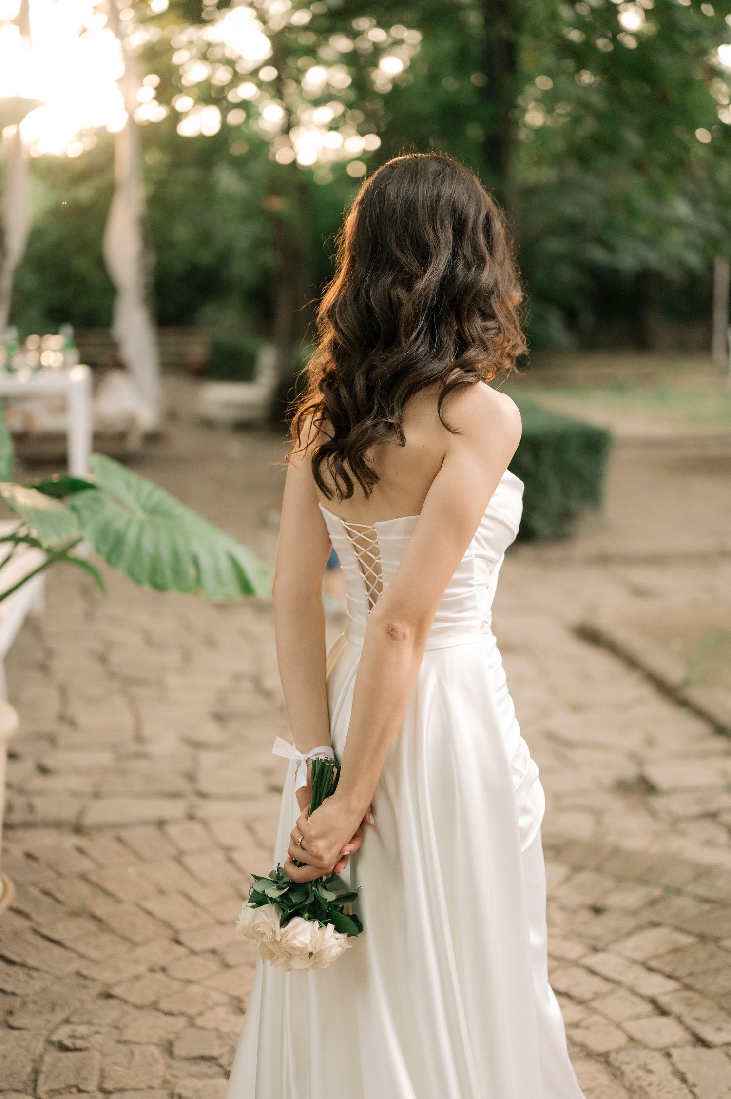 A bride in a white lace-up wedding dress holds a bouquet behind her back while standing on a stone path at sunset.