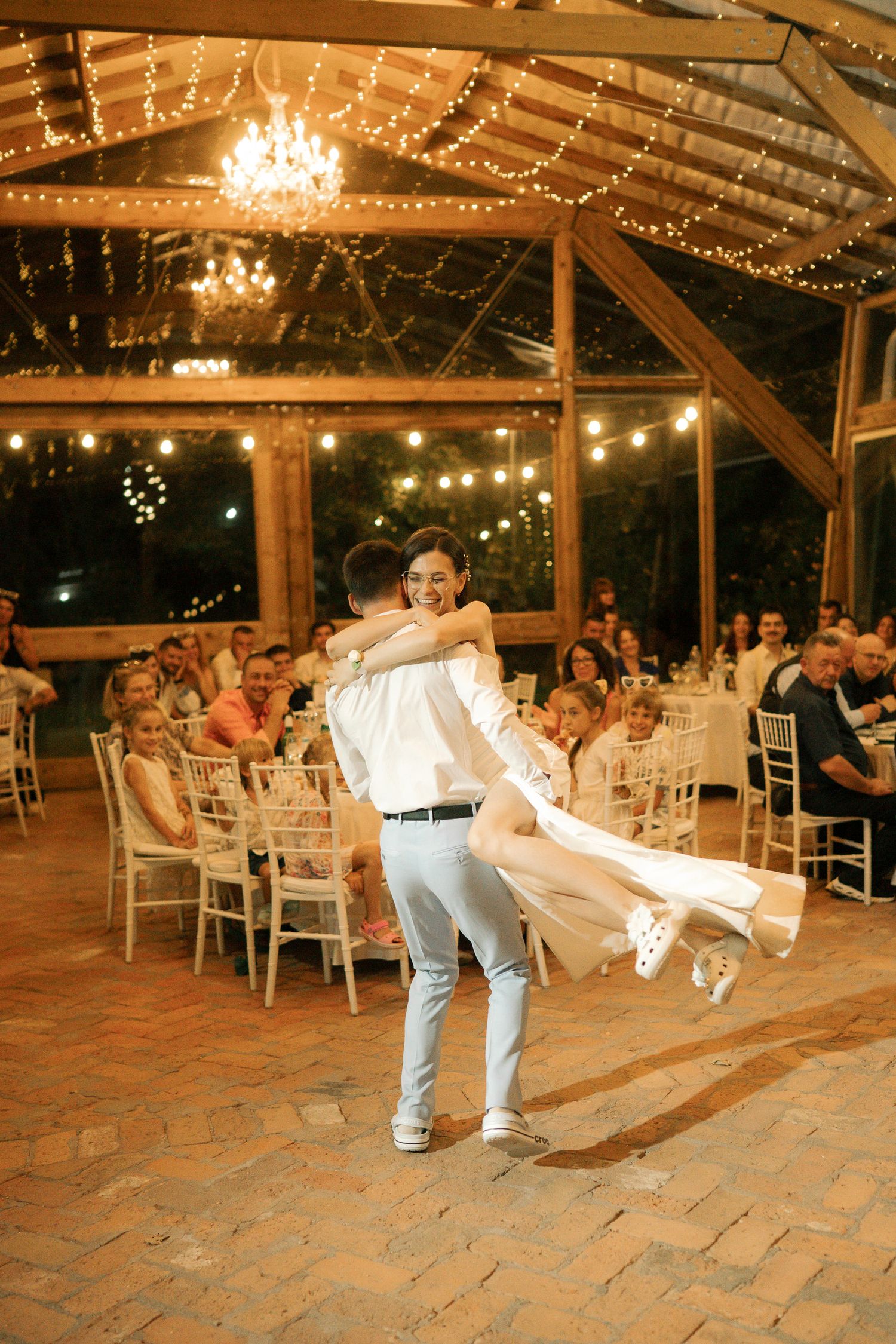 A couple performs a dip dance move during a reception in a rustic barn venue decorated with twinkling string lights.
