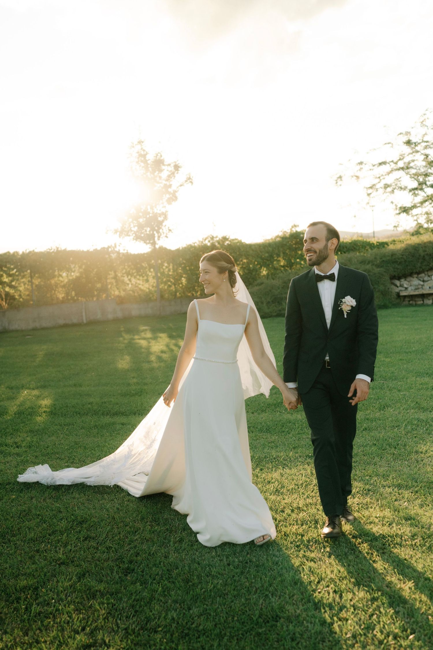 A couple in wedding attire walks together on a grassy lawn during golden hour sunset.