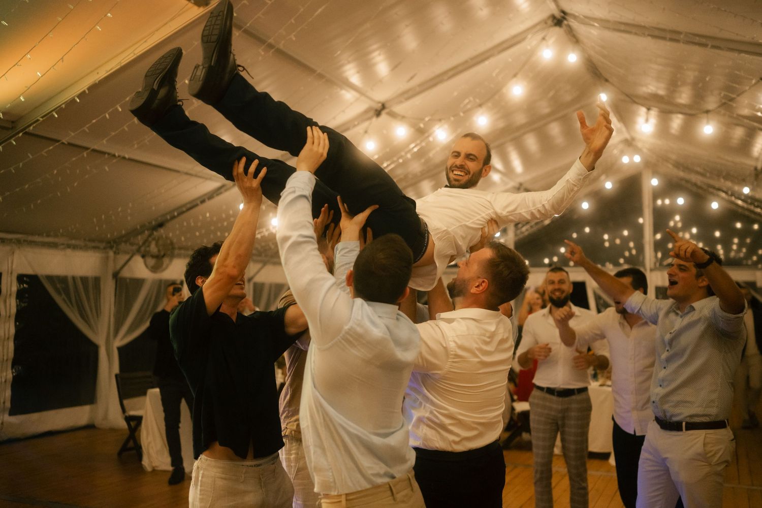 Wedding guests dance together under a white tent decorated with string lights during an evening reception celebration.