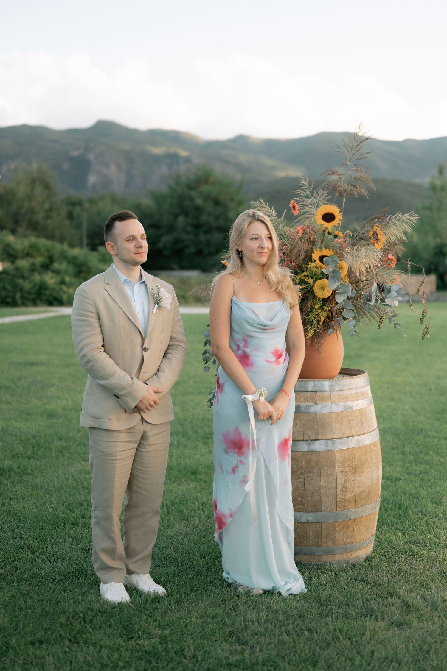 A couple poses outdoors in formal attire next to a rustic wine barrel decorated with sunflowers in a scenic mountain setting.