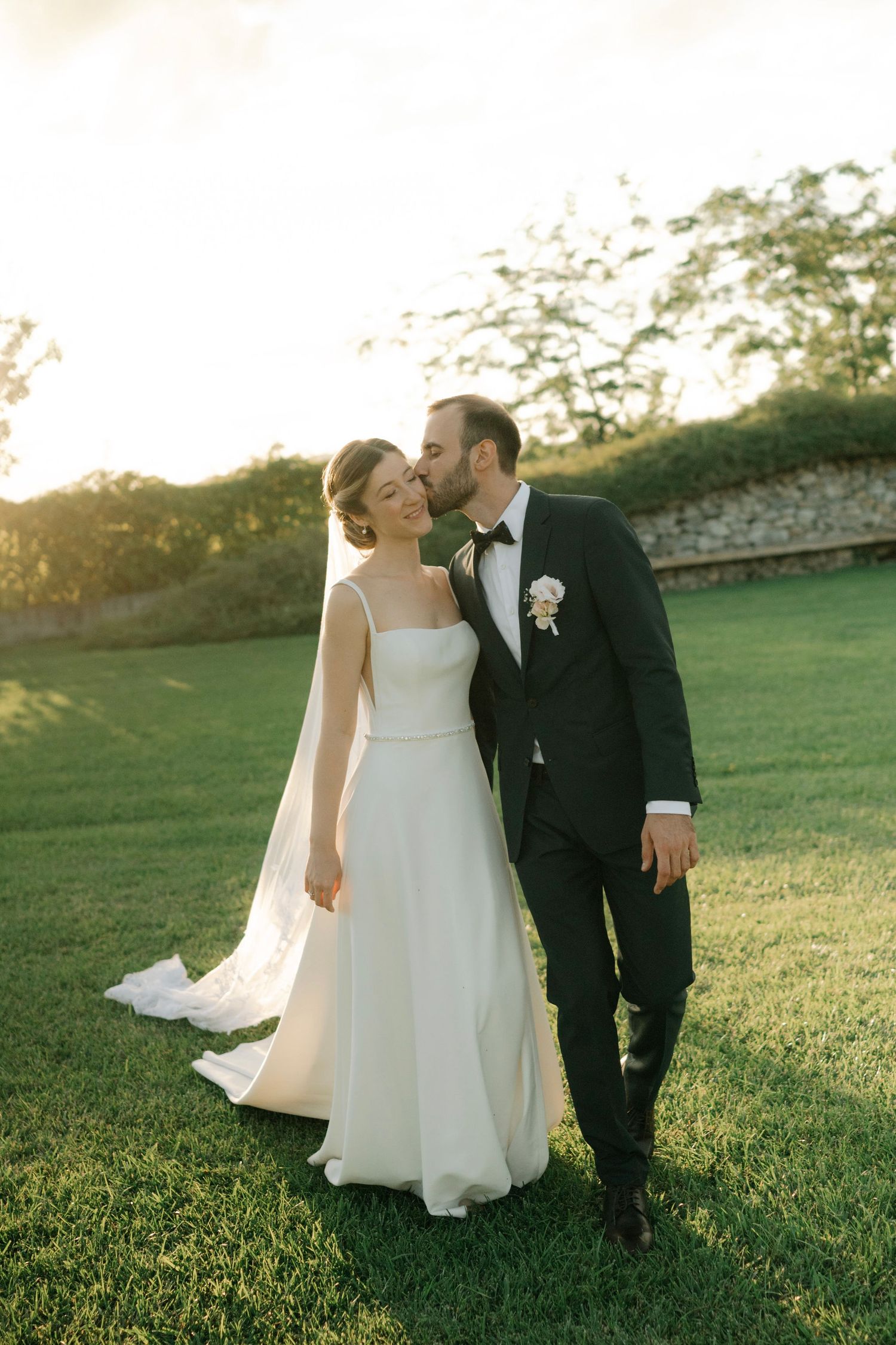 A romantic couple in wedding attire shares tender moments on a grassy hillside at sunset.