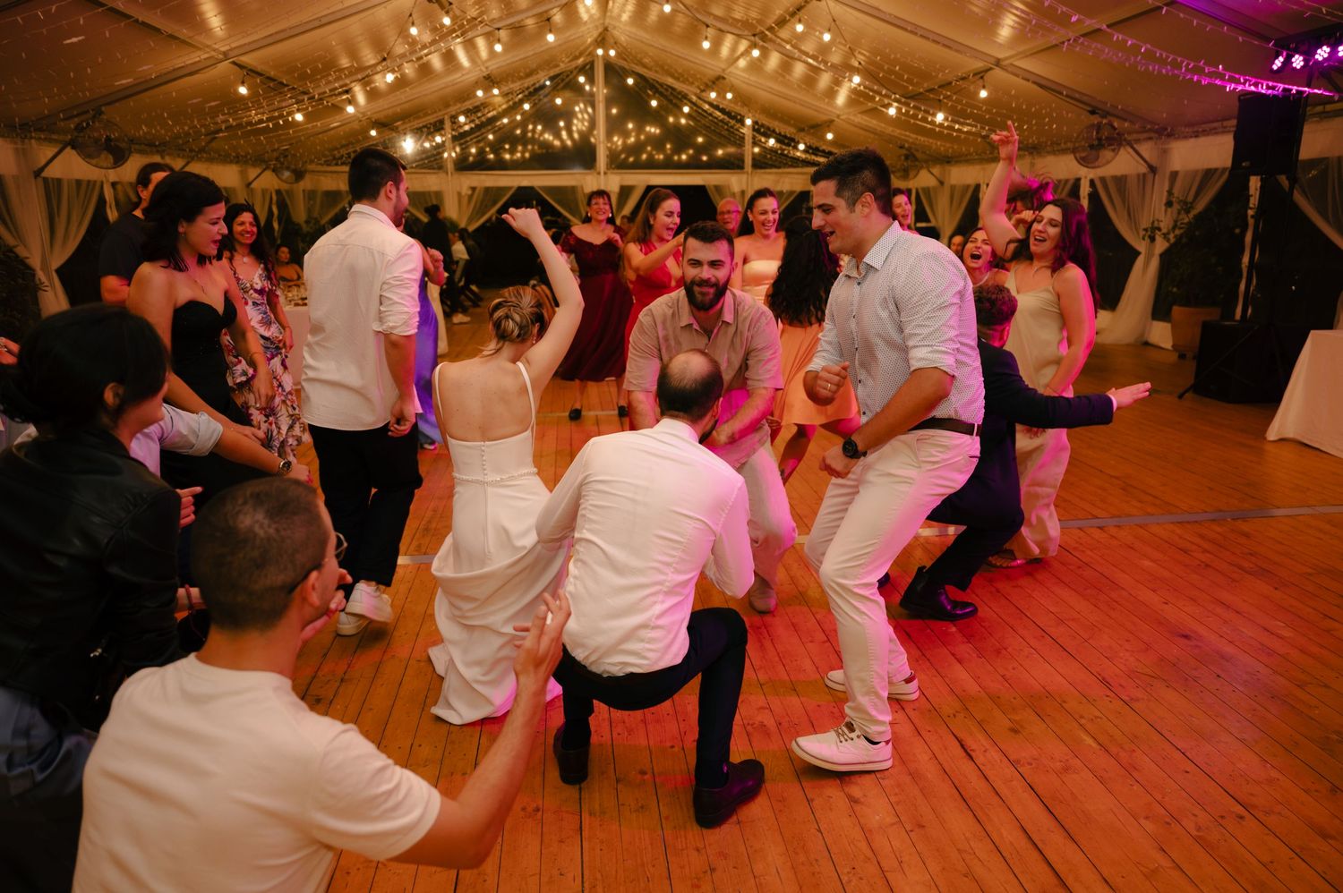 Wedding party dancing on a wooden floor under warm lighting in a decorated reception tent.