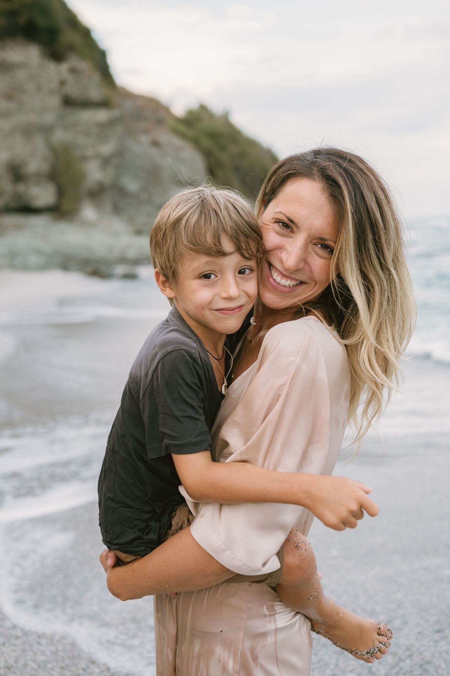 A smiling woman holds a young boy on a sandy beach with rocky cliffs and ocean waves in the background.