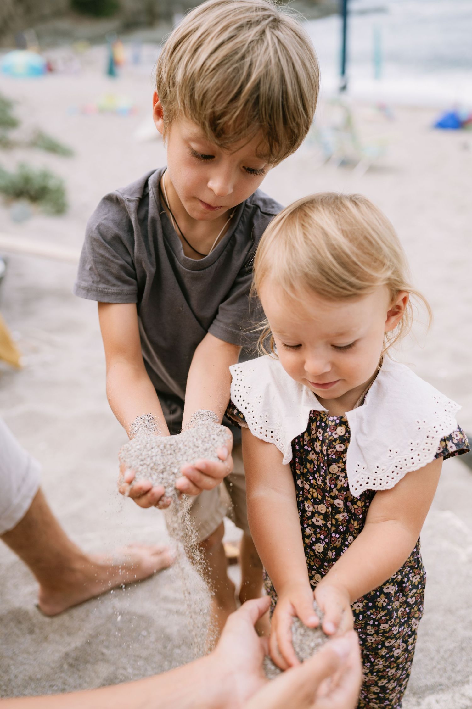 Two young children playing with sand on a beach, with an adult's hands visible helping them explore the sandy texture.