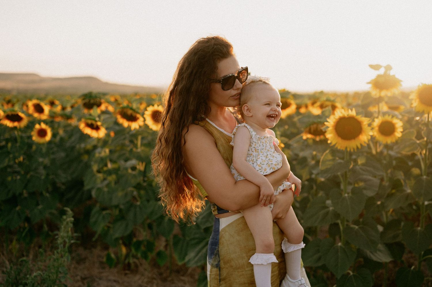 Mother holding laughing baby in sunflower field at golden hour sunset, both smiling joyfully outdoors.
