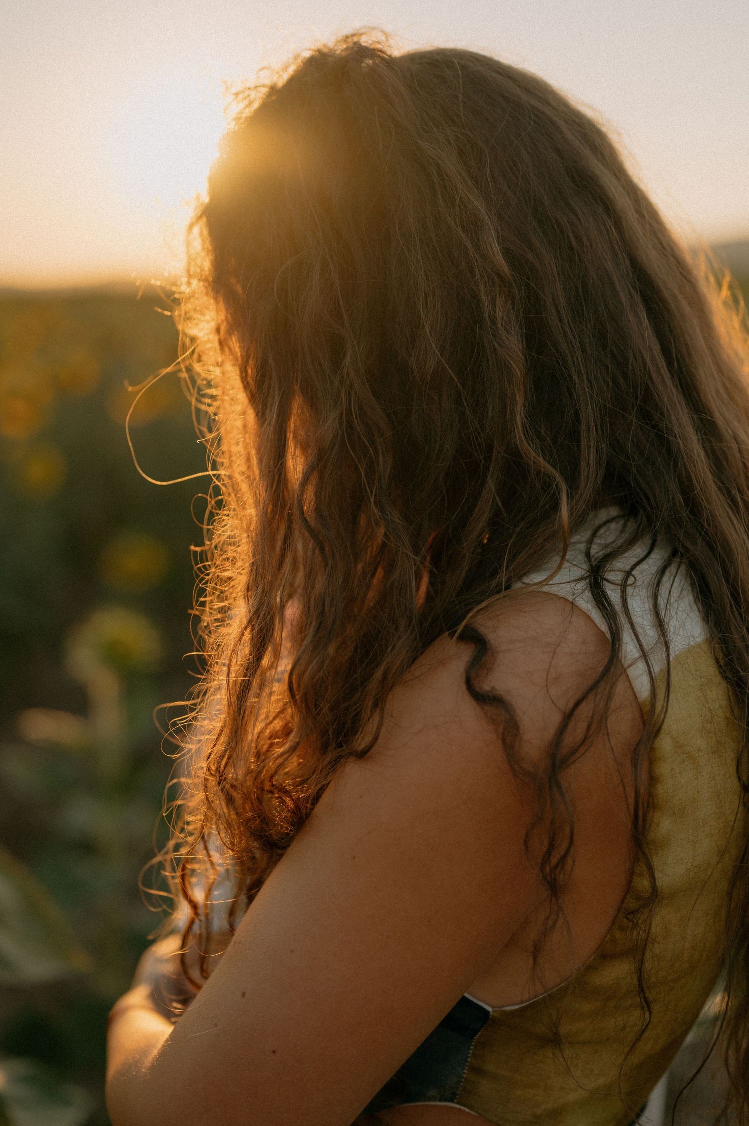 Woman with long wavy hair backlit by golden sunset light in a sunflower field, wearing a yellow top.