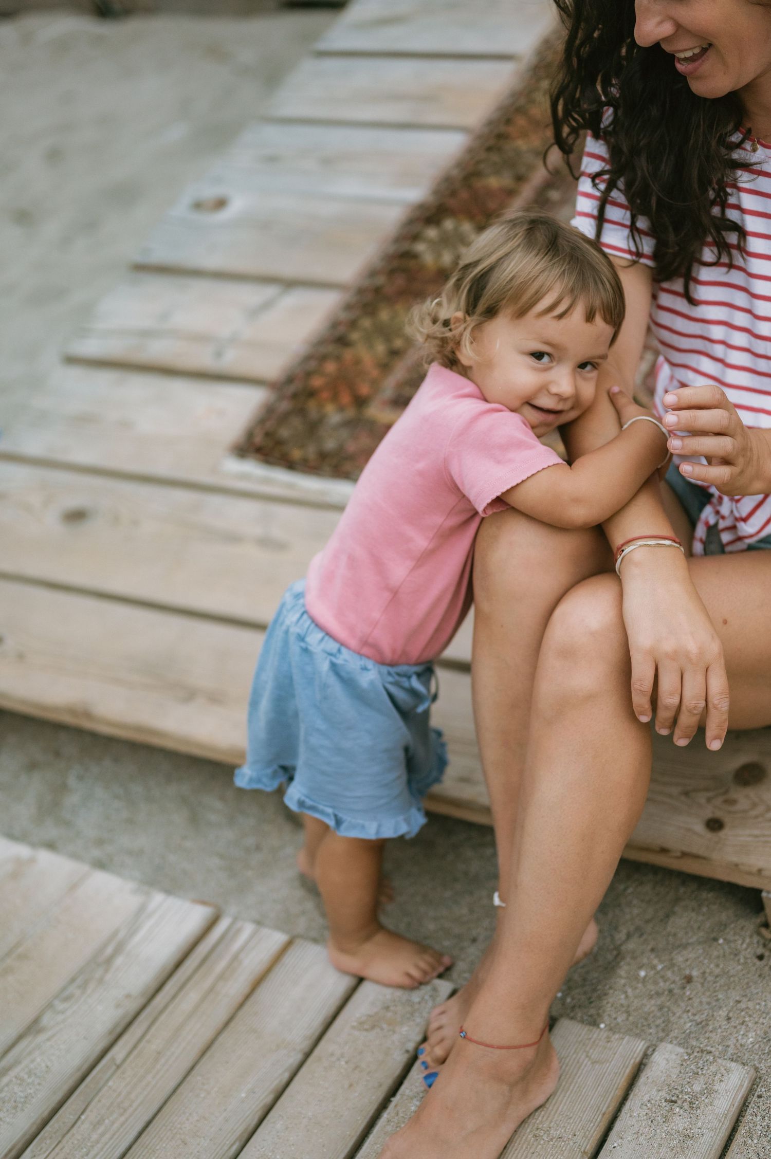A toddler girl in pink shirt and blue skirt hugs her mother's legs on wooden beach steps.
