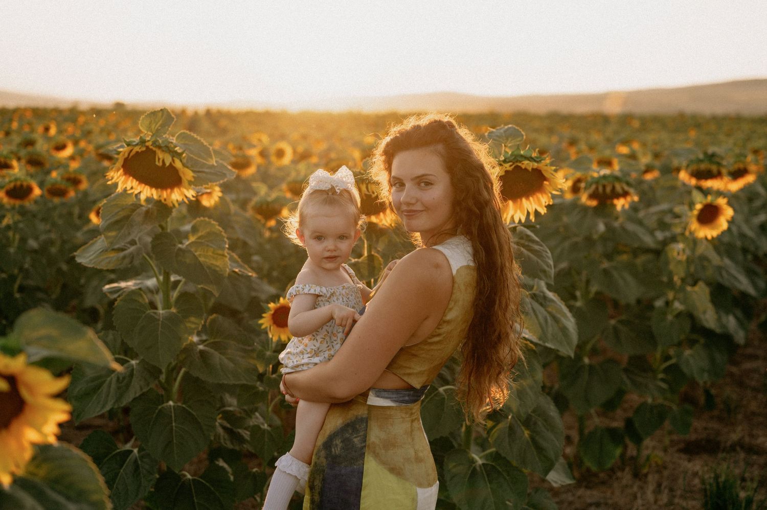 A young mother holds her toddler in a golden sunflower field at sunset, both smiling warmly.