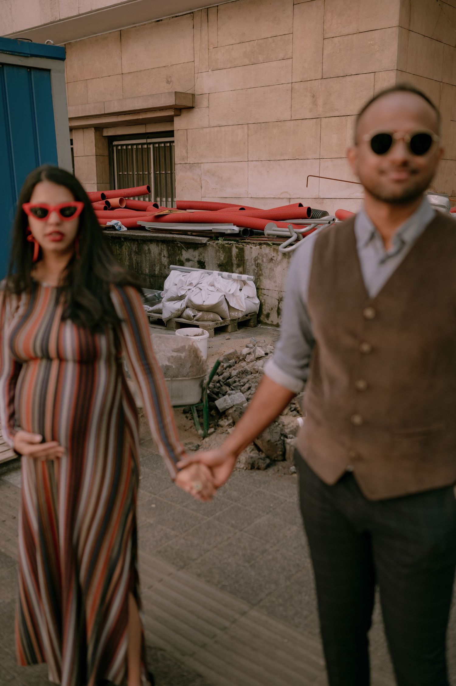 A stylish couple holding hands outdoors, woman in striped dress with red sunglasses, man in grey vest with sunglasses.