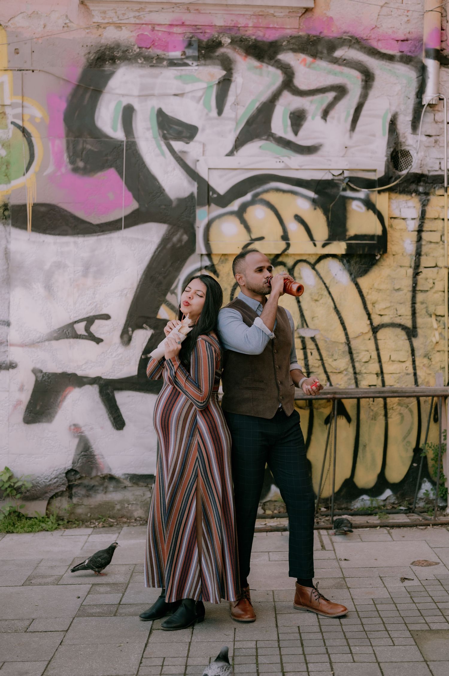 Stylish couple posing back-to-back in front of colorful graffiti wall, woman in striped dress, man in vest.
