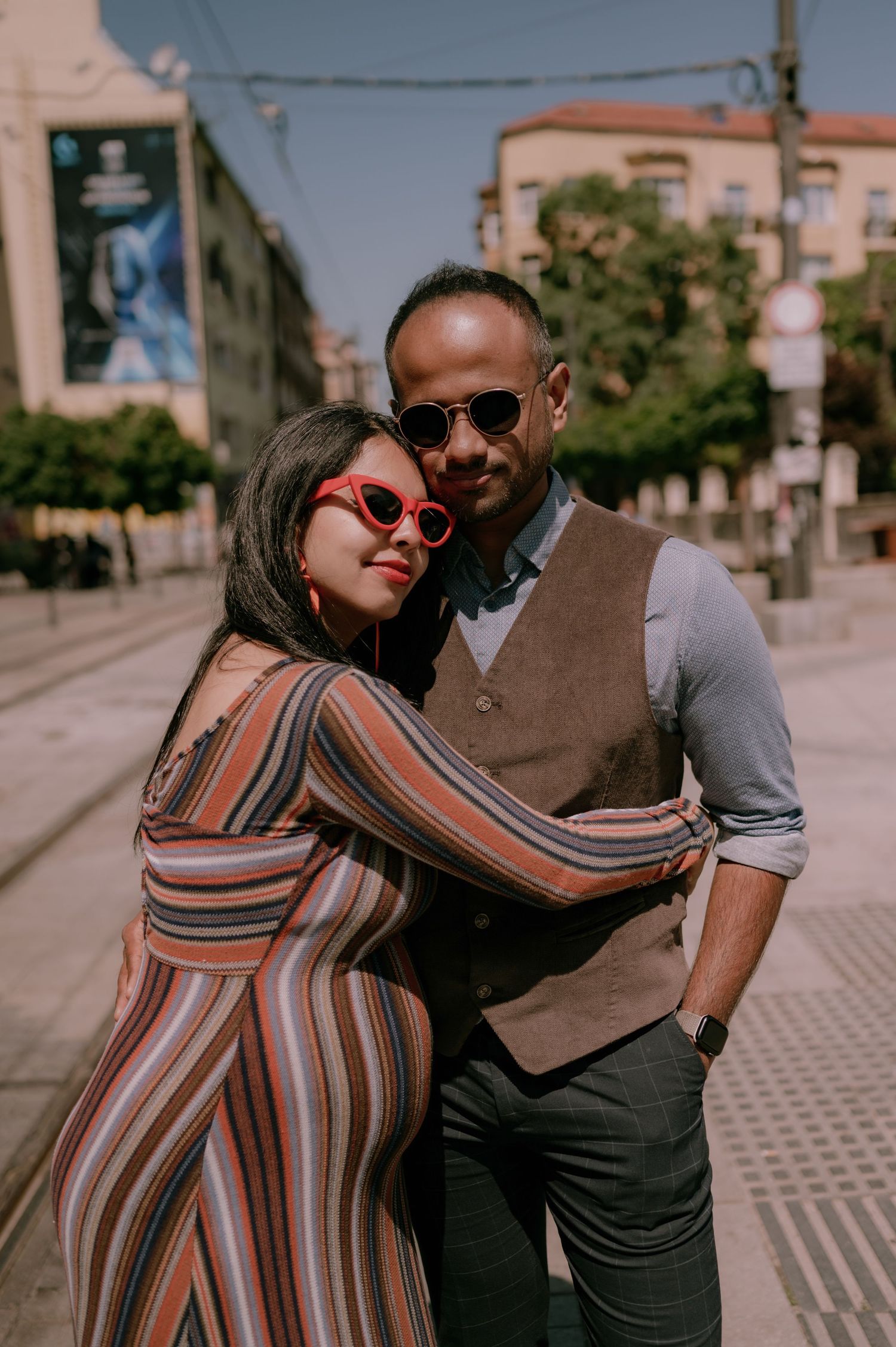 Stylish couple posing on a sunny urban street, woman in striped jumpsuit with red sunglasses leaning on man in vest.