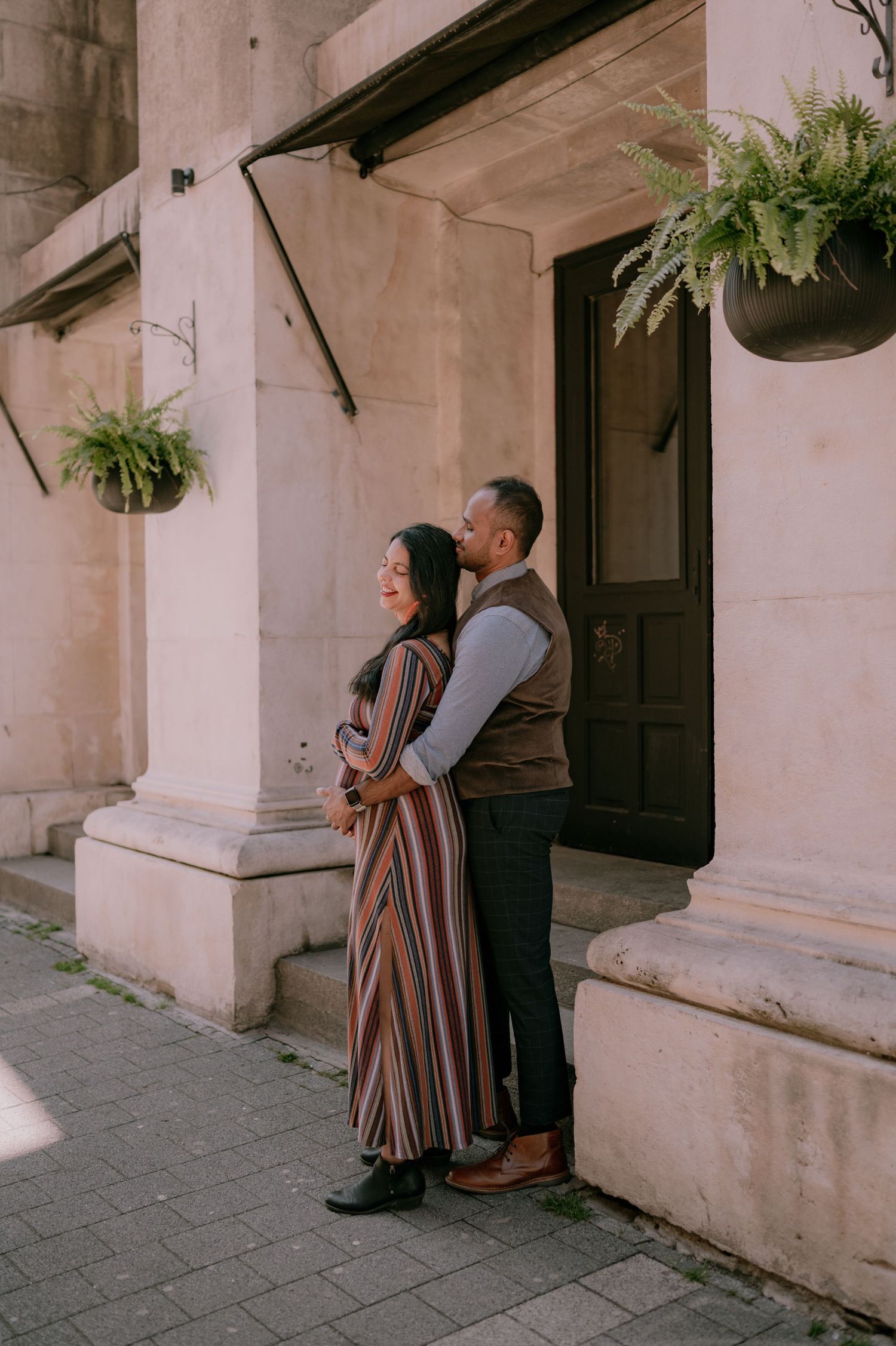 Couple embracing outside a historic building with hanging fern planters, woman in striped maxi dress, man in vest.