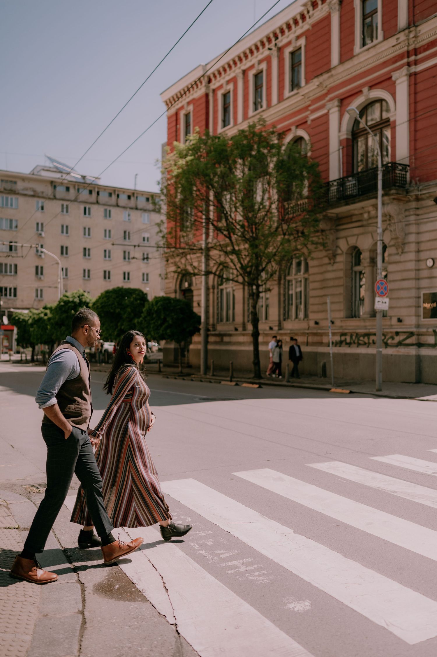 Couple crossing a zebra crossing in an urban street with a red historic building and trees in the background.