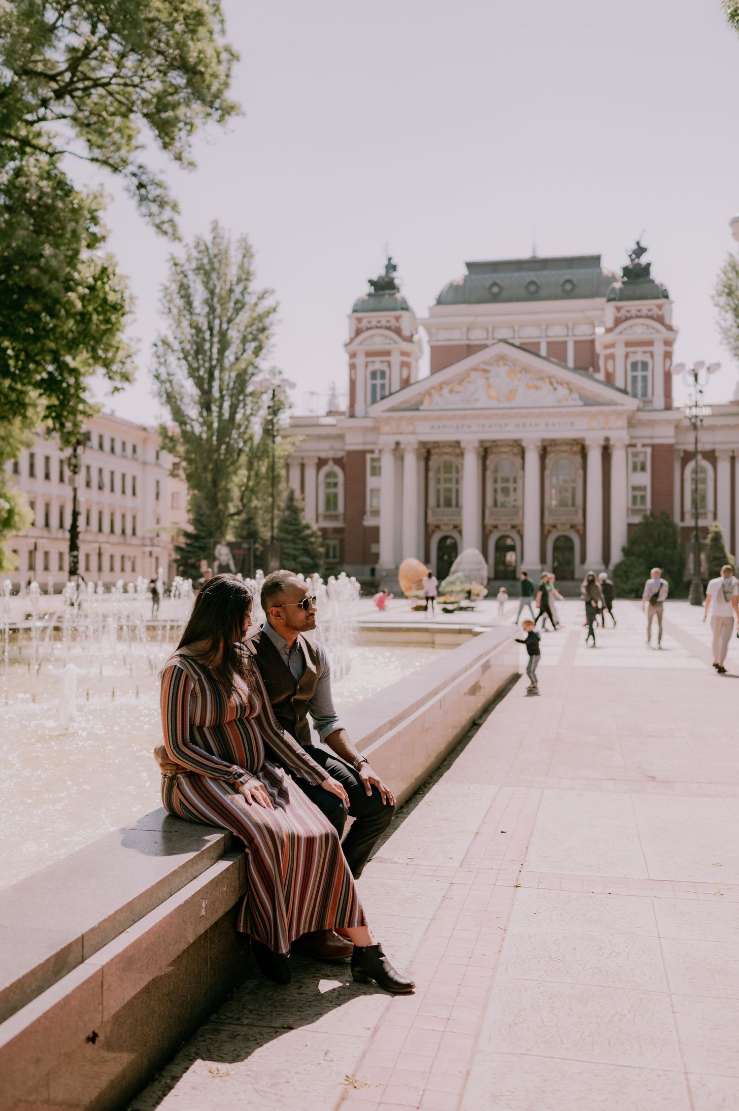 Couple sitting by a fountain in a sunny plaza with a grand neoclassical building in the background.