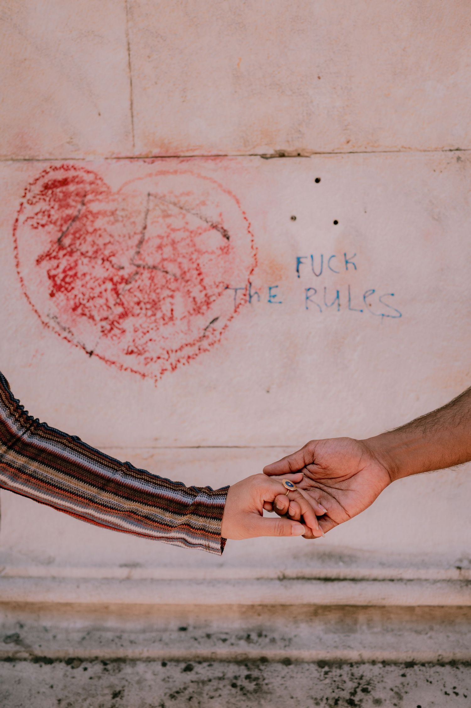 Two hands holding gently against a wall with a red heart graffiti and 'Fuck The Rules' text.