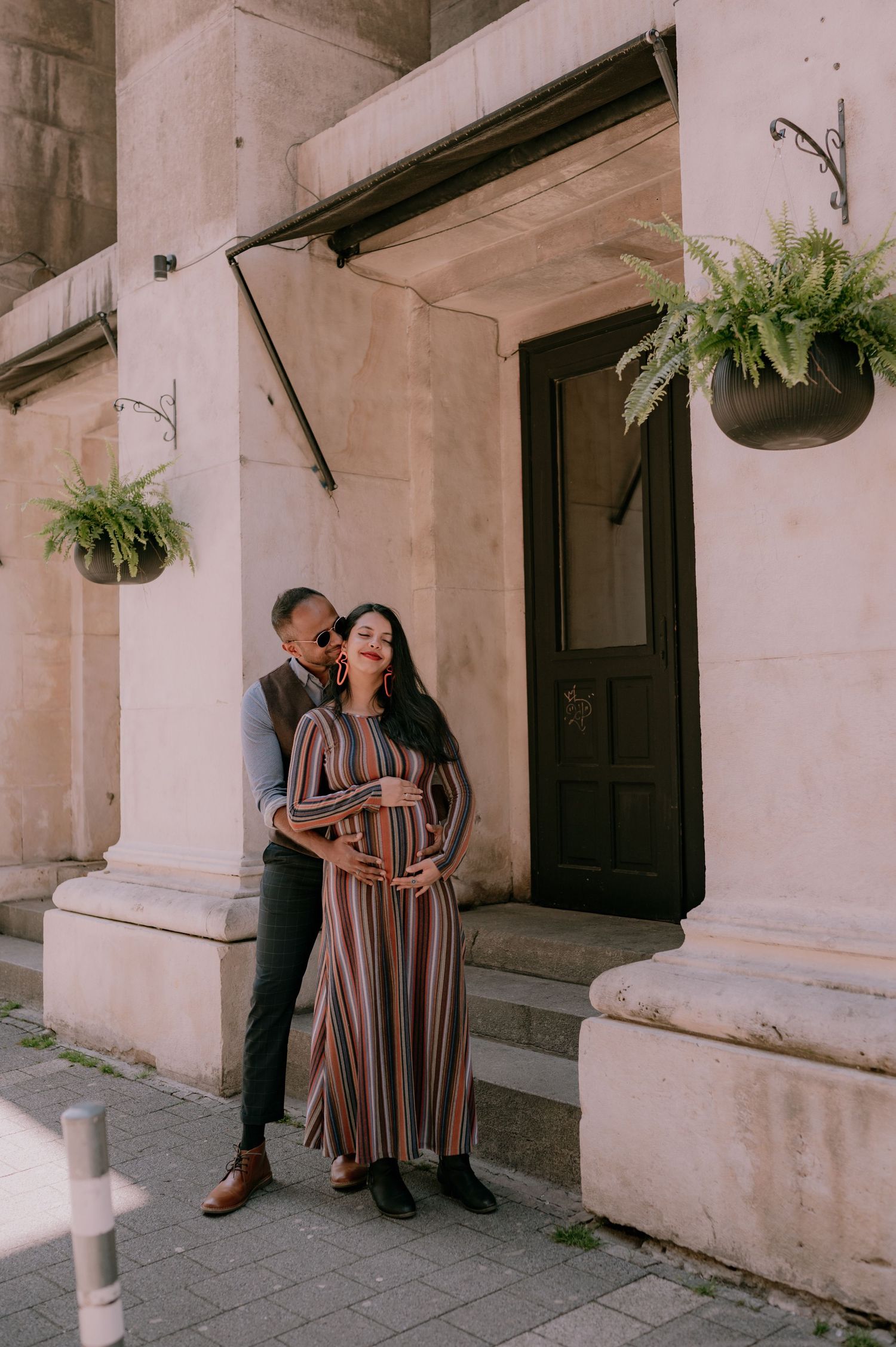 Couple posing outside historic building, woman in striped maxi dress cradling baby bump, man embracing her from behind.