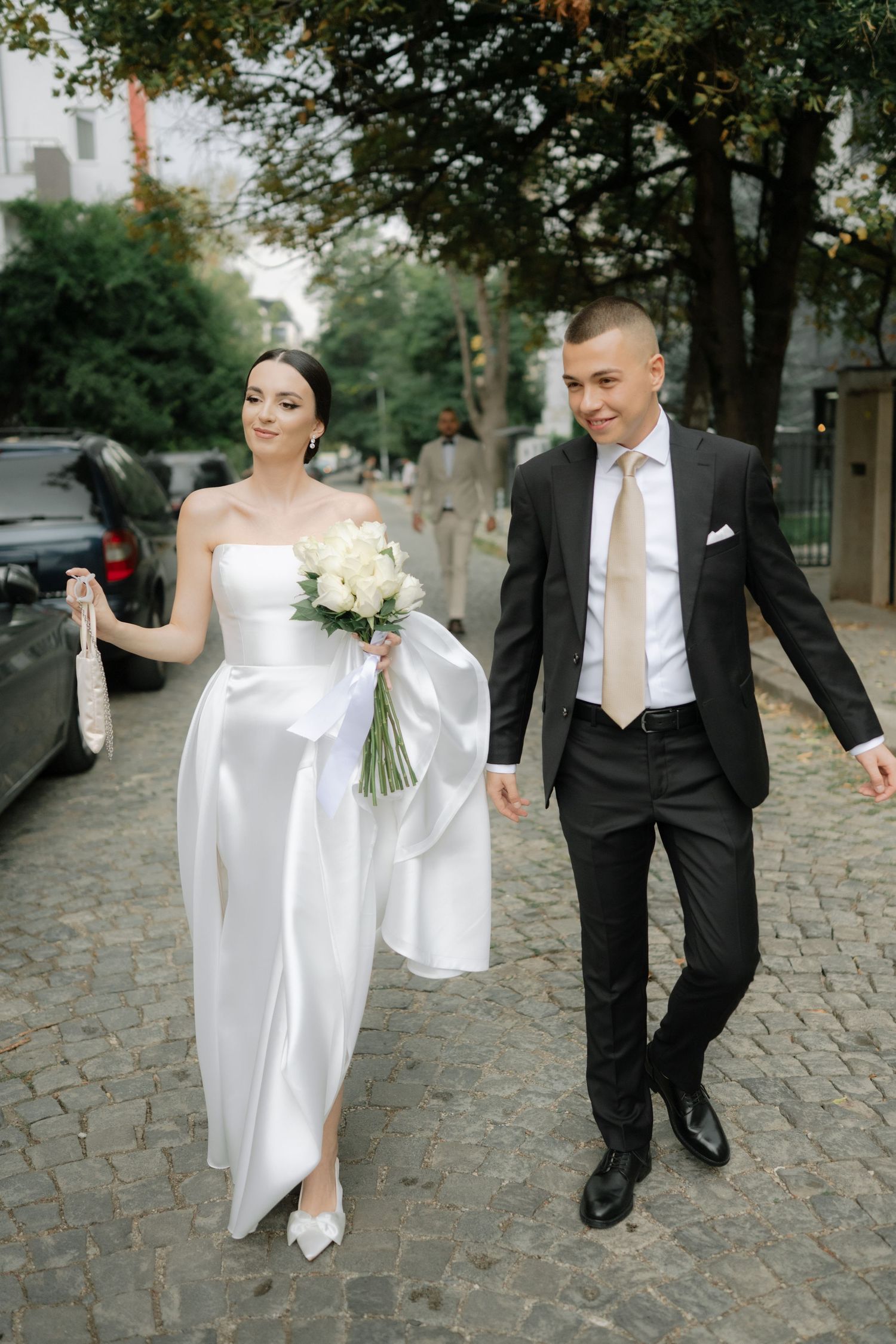 A couple in wedding attire walks together on a cobblestone street, with trees and buildings in the background.