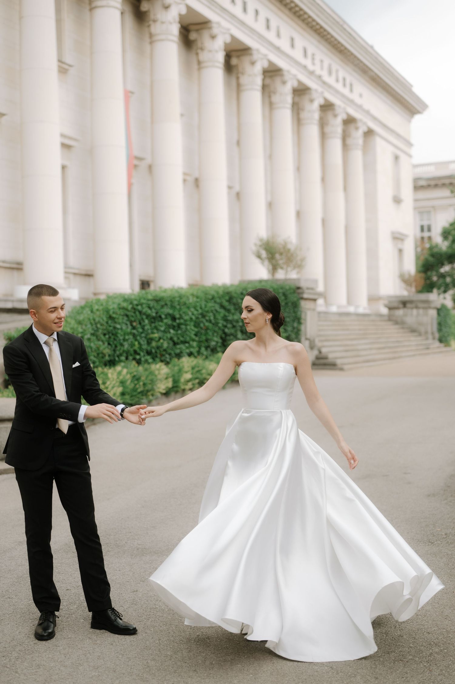 A couple in formal wedding attire twirls outside a grand neoclassical building with white columns.