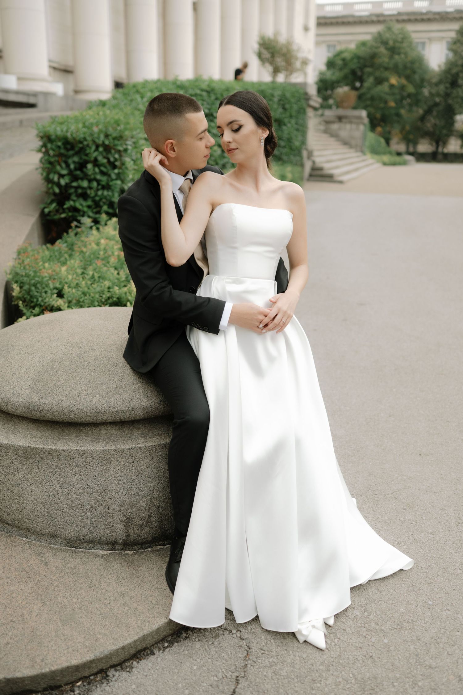 A couple in wedding attire share an intimate moment while sitting on a stone pillar outside a classic building.