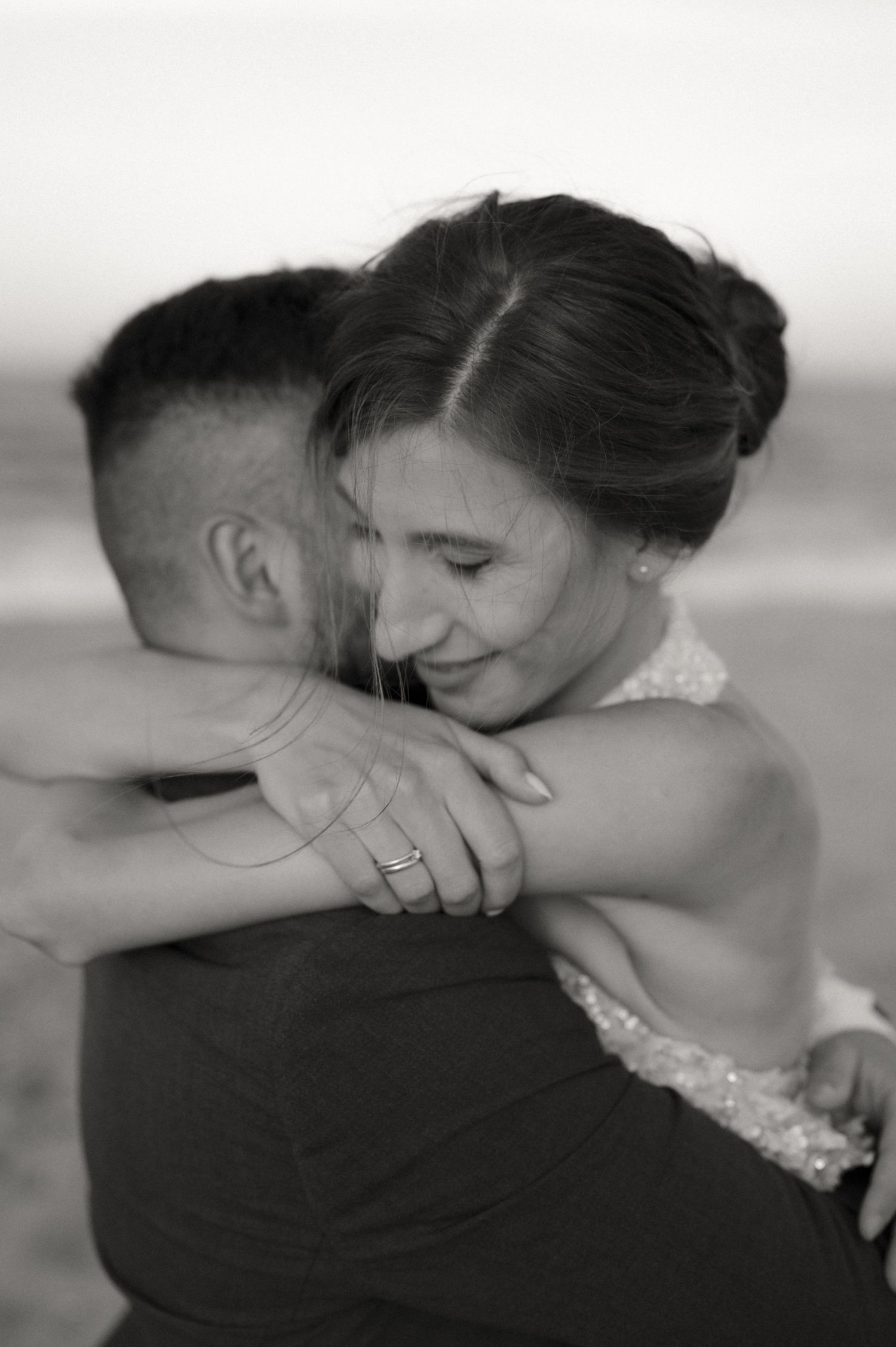 Black and white photo of a smiling bride hugging her groom on a beach, wearing a wedding ring.
