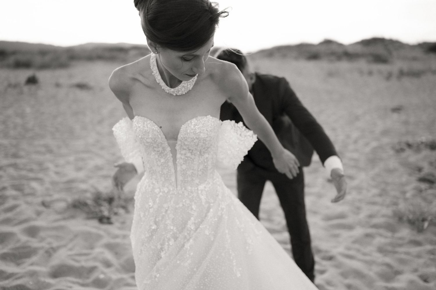 Black and white photo of a bride in a beaded strapless gown and groom in a suit on a sandy beach.