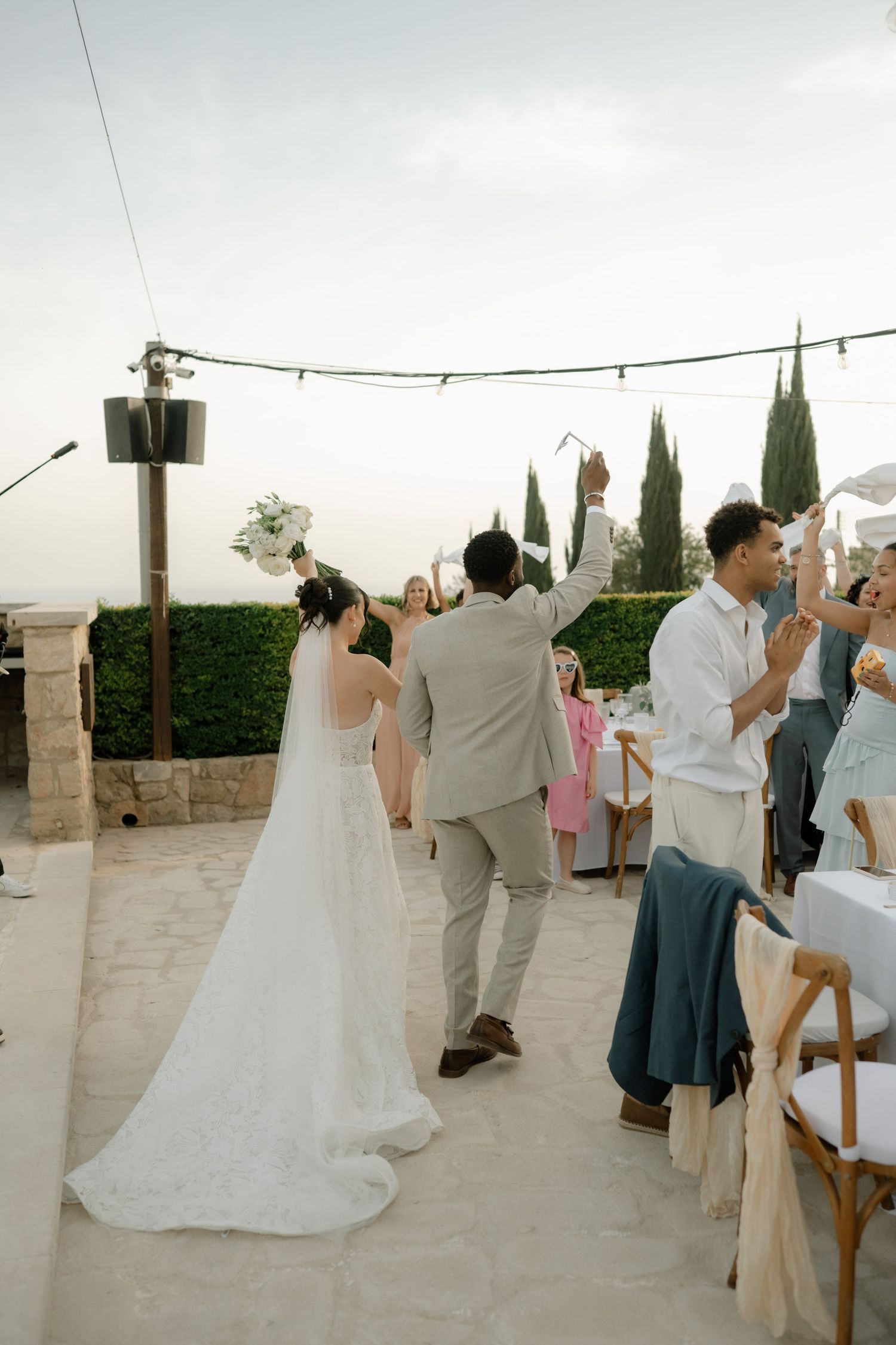 Bride and groom walk hand-in-hand at outdoor wedding reception as guests celebrate under string lights.