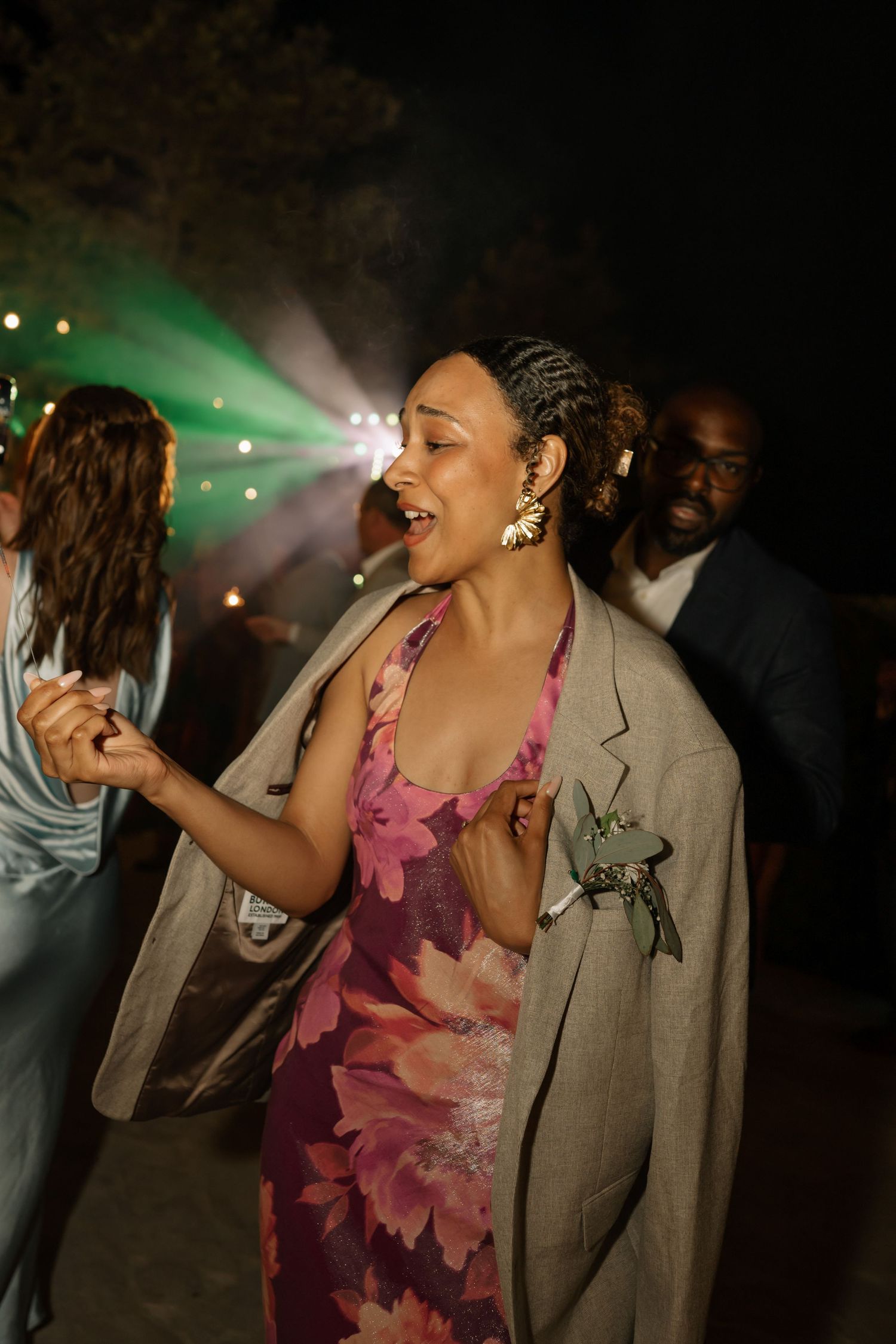 Woman in floral dress and blazer dancing joyfully at an outdoor evening party with colorful lights.