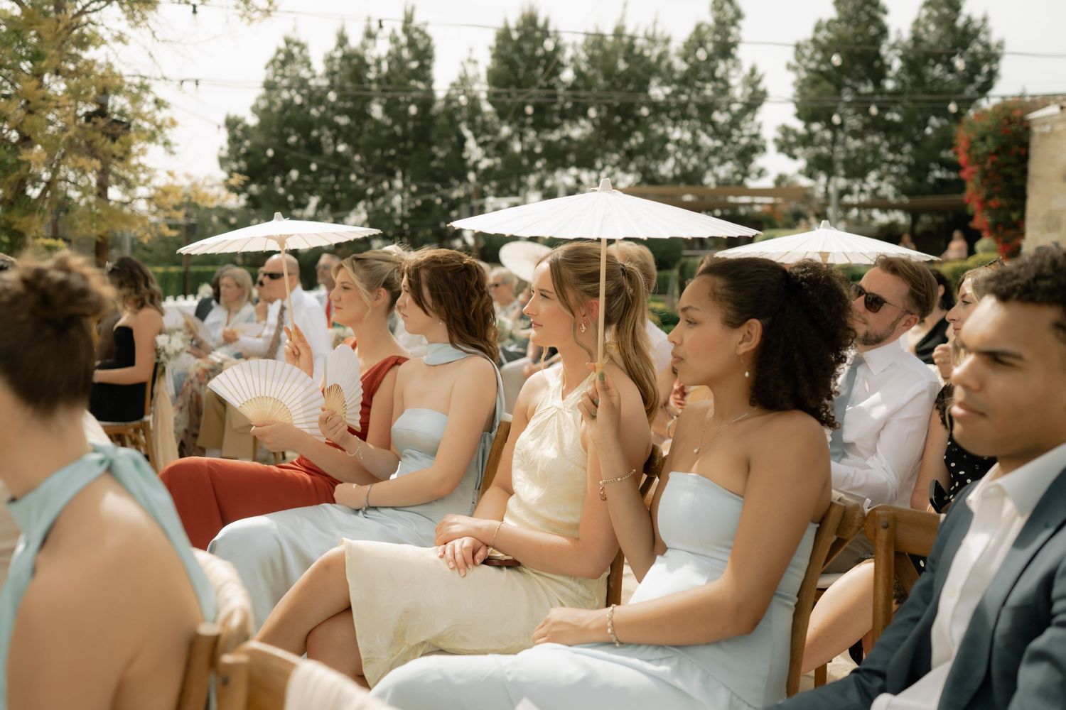 Guests seated outdoors at a wedding ceremony, wearing elegant formal attire on a sunny day.