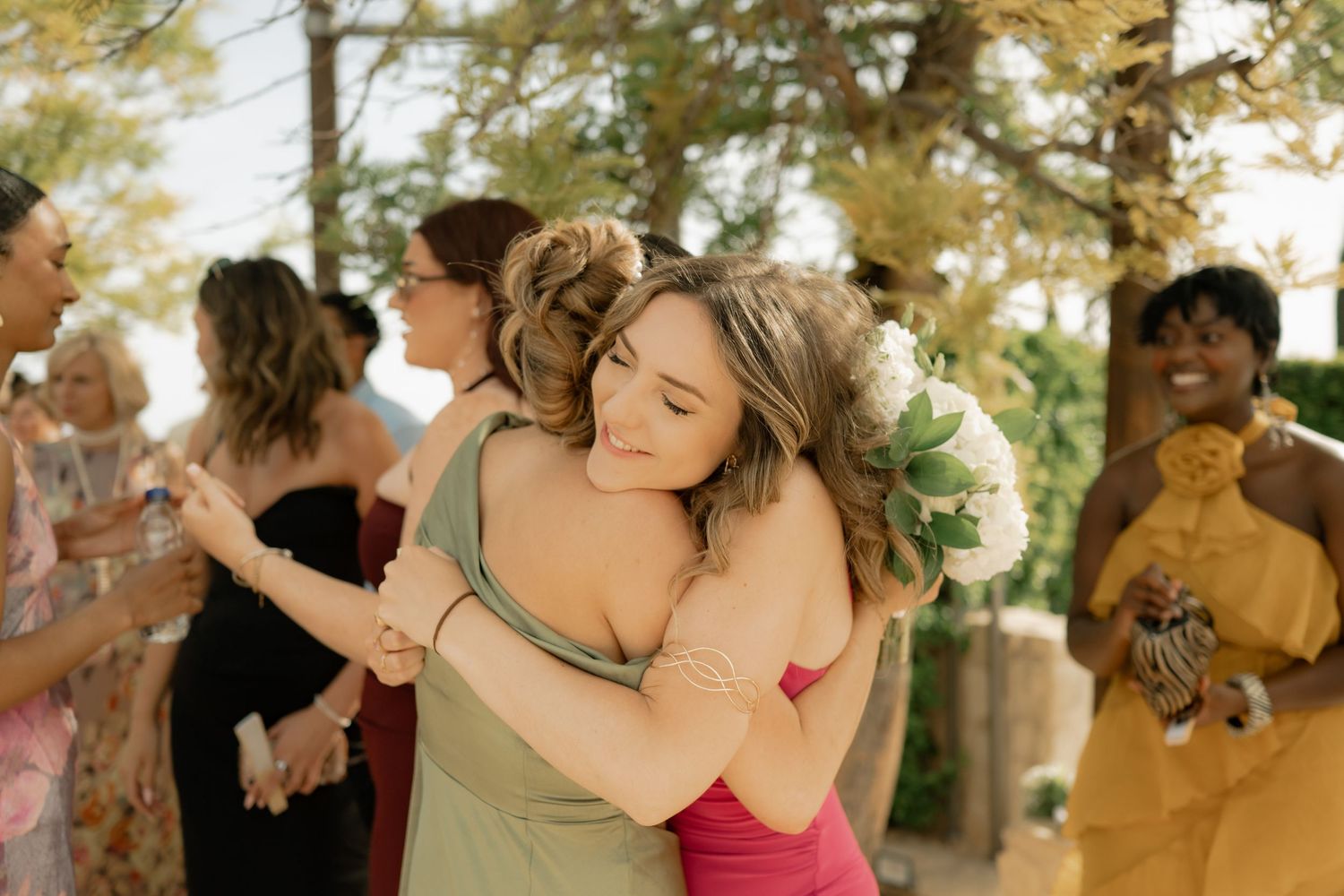 Two women share a warm hug at a wedding celebration, one holding a bouquet, surrounded by guests outdoors.