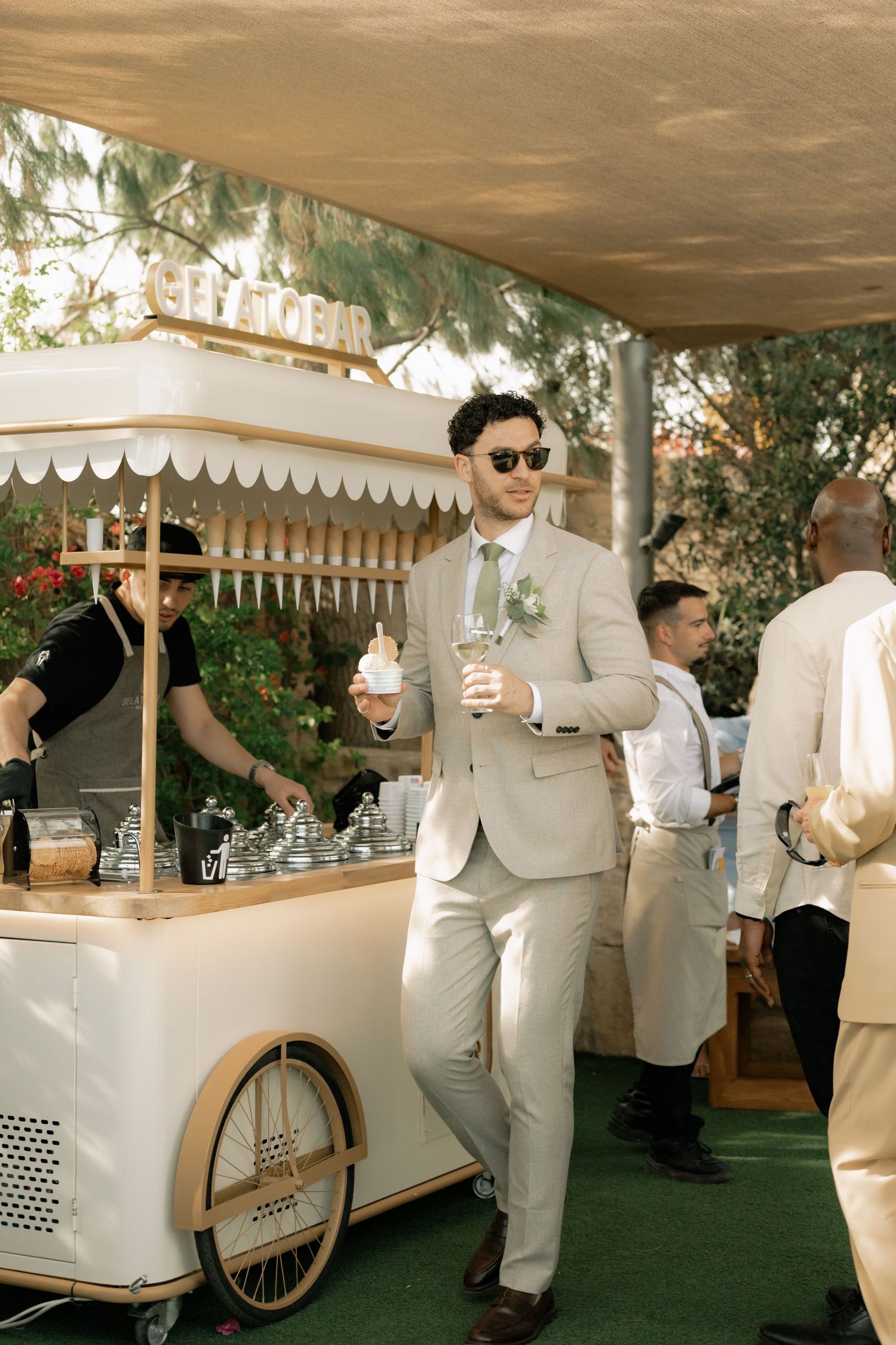 Man in beige suit with sunglasses stands by elegant white gelato bar cart at outdoor wedding reception.
