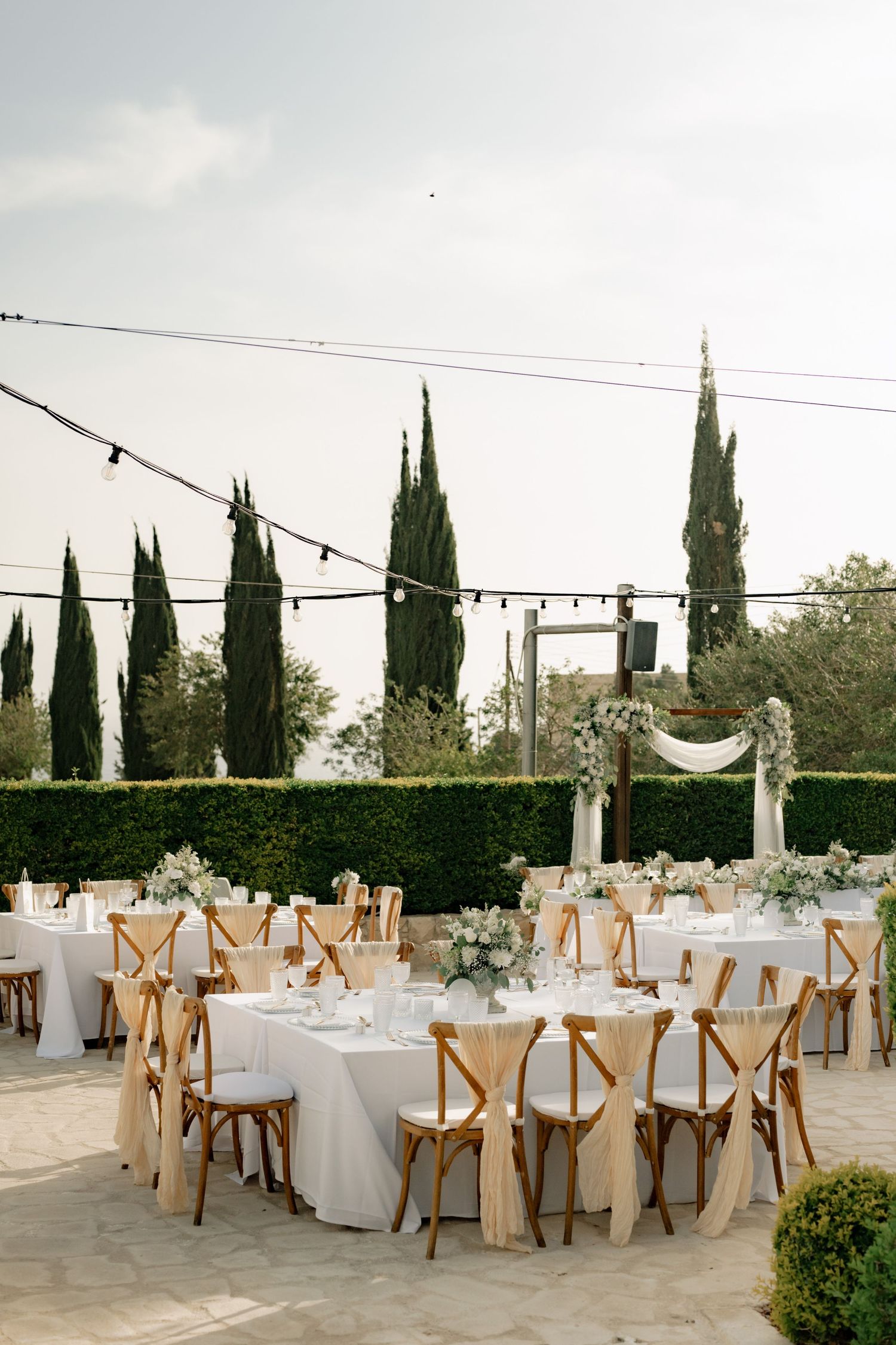 Elegant outdoor wedding reception with round tables, wooden cross-back chairs, white linens, floral centerpieces, and cypress trees.