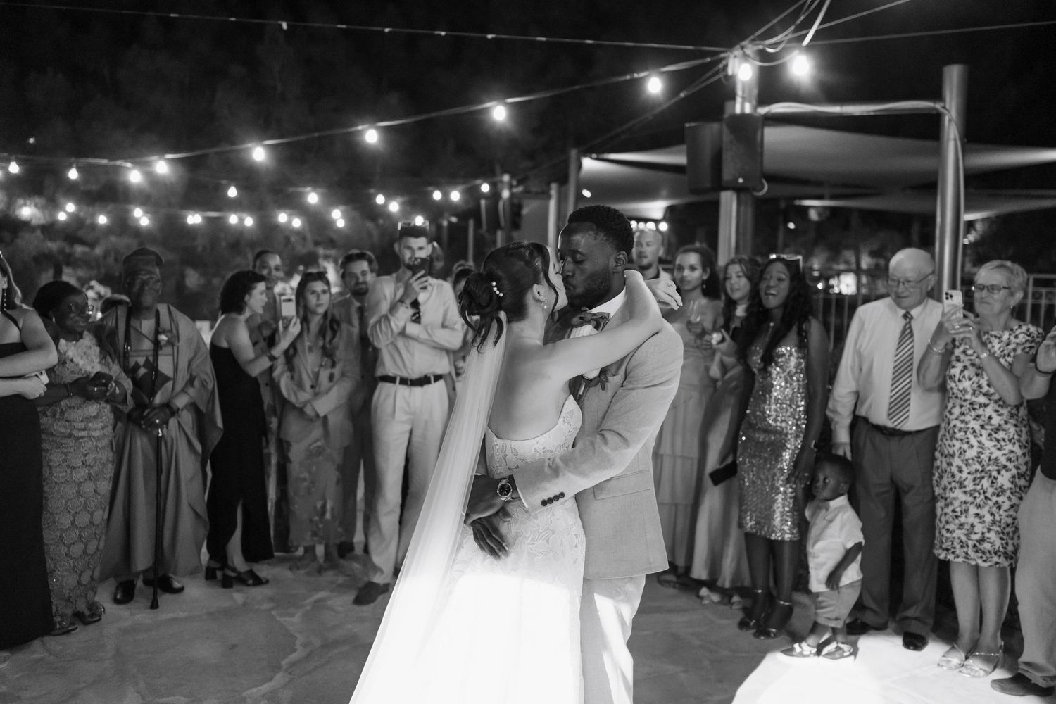 Bride and groom share their first dance surrounded by guests under string lights at an outdoor wedding reception.