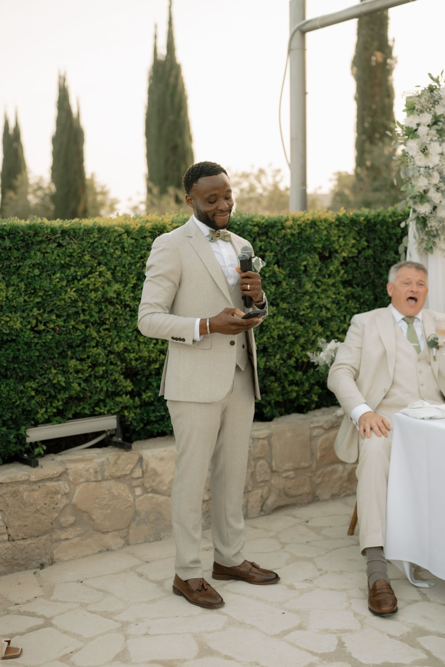 A groom in a beige suit gives a speech at an outdoor wedding reception while an older guest laughs in the background.