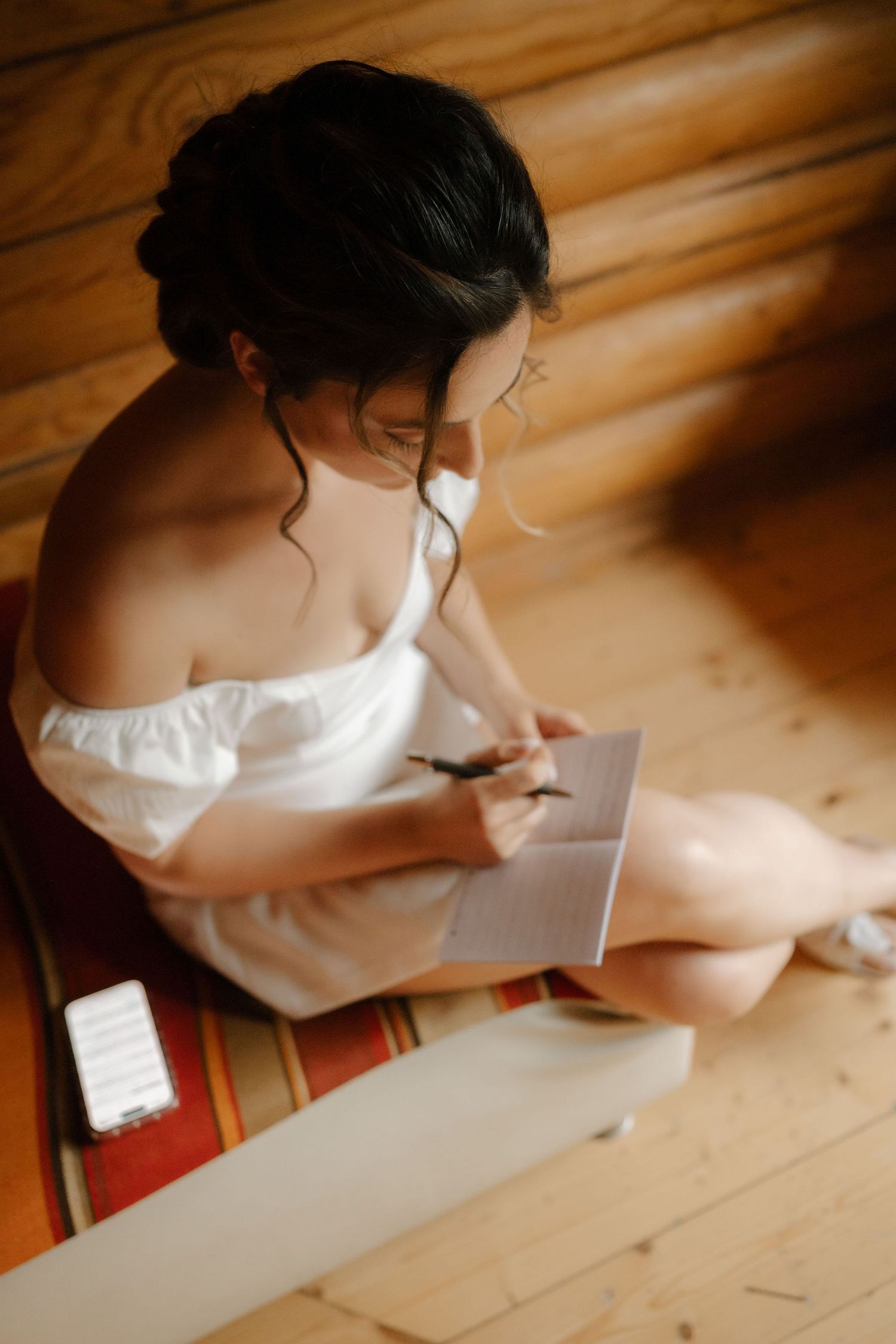 Person in white dress writing wedding vows on paper while sitting at wooden table in soft natural light.